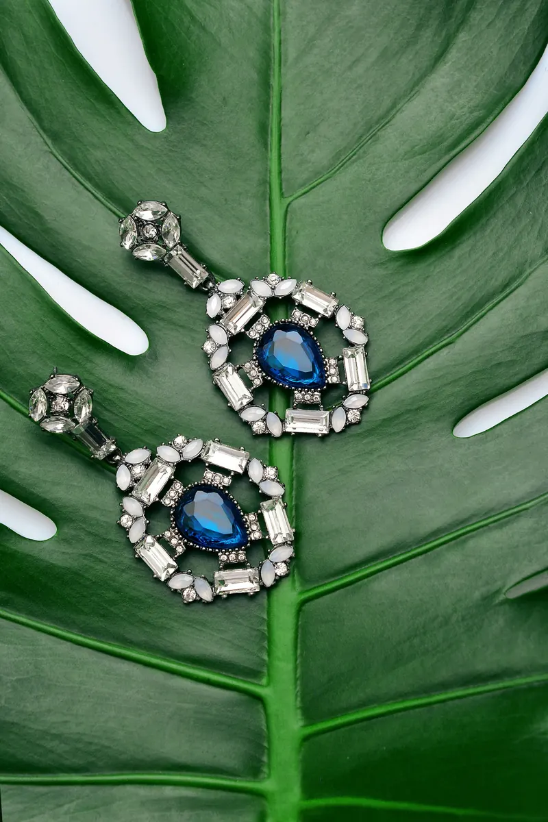 Ornate blue-stone and crystal drop earrings on a green monstera leaf, the kind of sapphire-toned piece that suits a Skyrim earrings look