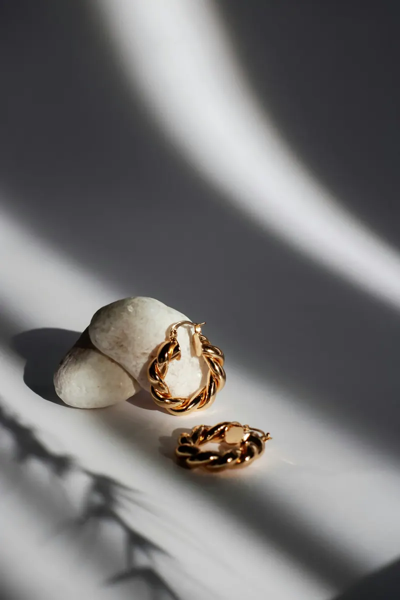 Twisted gold hoop earrings on white pebbles with dramatic shadows, clean and polished